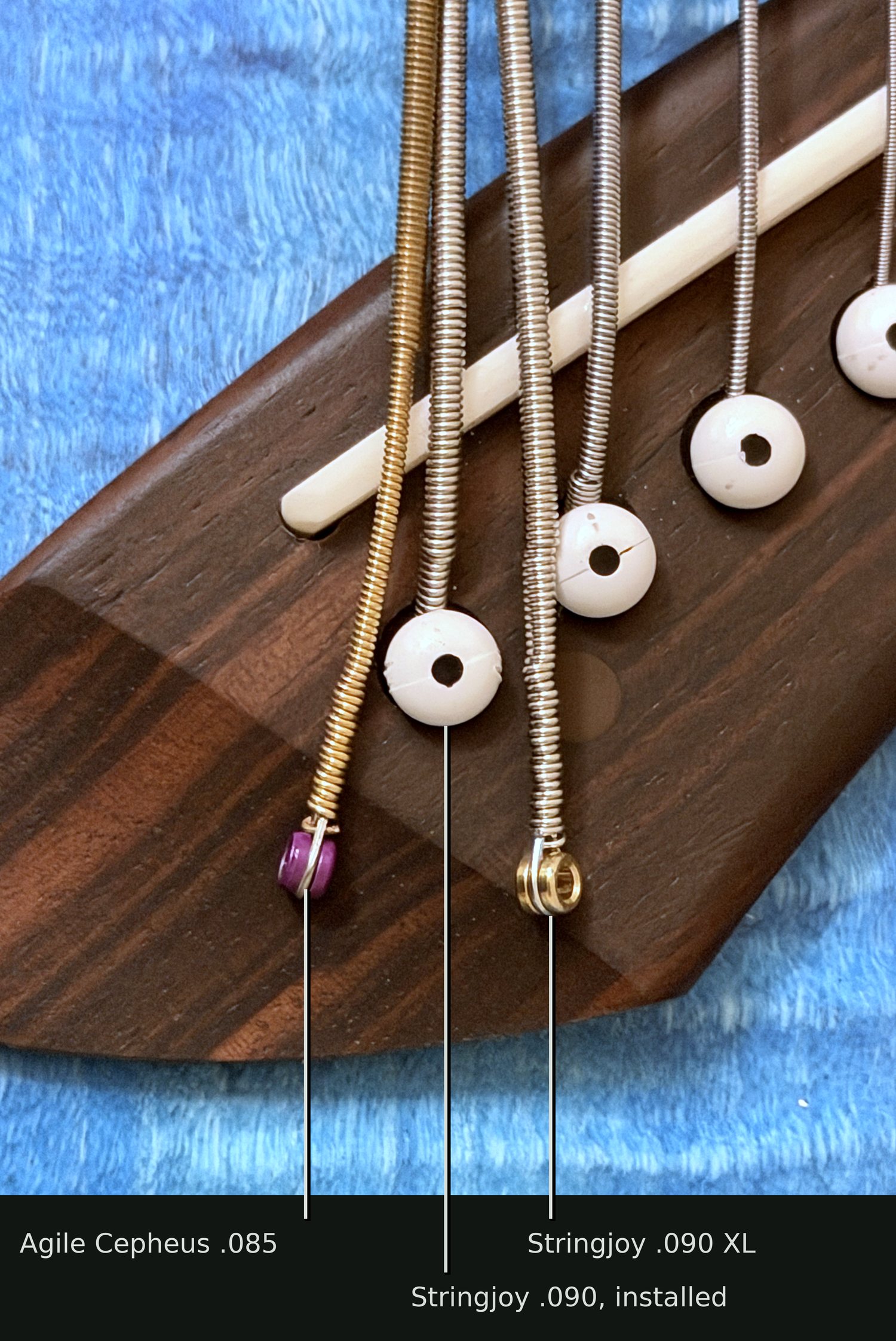 The dark wooden bridge of an 8-string acoustic guitar, with white bridge pins. In the middle, a Stringjoy .090 is installed, held in its hole by a pin. Lying loose to its left is the gold Agile Cepheus .085 with a purple ball end; lying loose to its right is the Stringjoy .090 XL with a brass ball end.