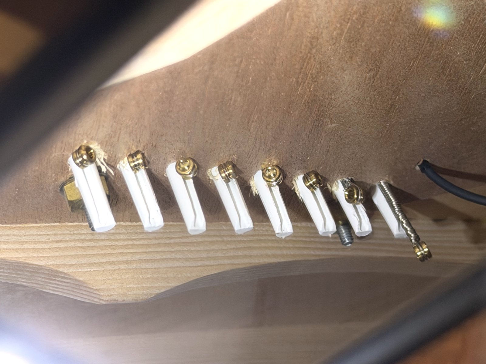 Underside of the guitar’s bridge plate, seen from inside the body: a row of eight white bridge pins, with brass string ball ends seated against the plate beside them. At the right end, the heavy Stringjoy .090 hangs loose beside its pin, its ball end dangling below instead of seated.