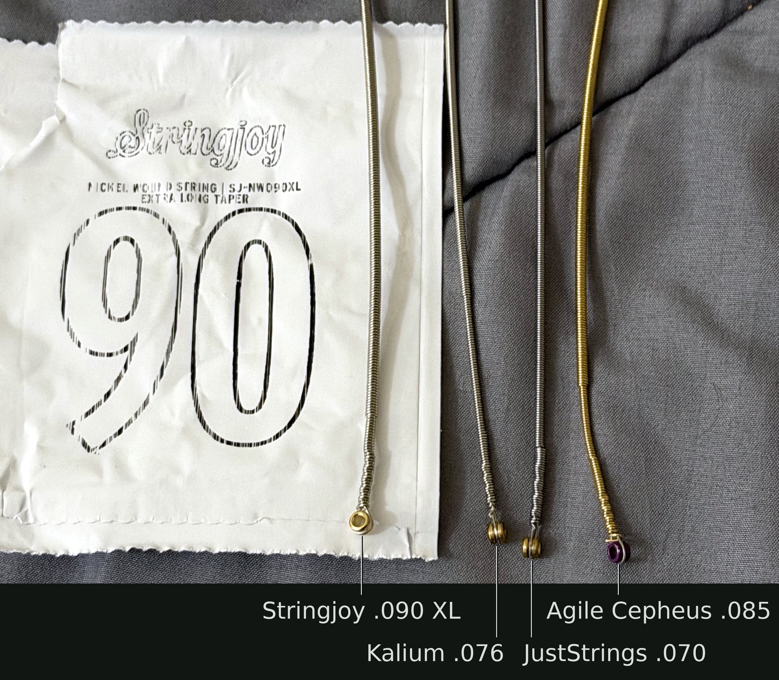 Four heavy wound guitar strings lying side by side, ball ends at the bottom. Far left, on its white Stringjoy packet printed “90” and “SJ-NW090XL Extra Long Taper”, is the Stringjoy .090 nickel-wound XL, with a brass ball end; its winding narrows slightly just above the ball end. Next is a Kalium .076, whose winding narrows a little further up, then a JustStrings .070, which doesn’t narrow at all. Far right is a gold-colored Agile Cepheus .085 with a purple ball end, whose winding narrows much more noticeably near the ball end.